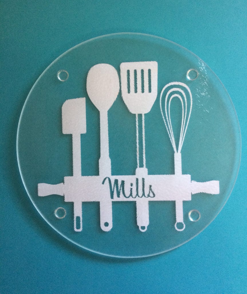 Round, clear glass acrylic with a white design. The design includes a rolling pin, spatula, spoon, spatula, and whisk, along with the name "Mills" written in cursive.