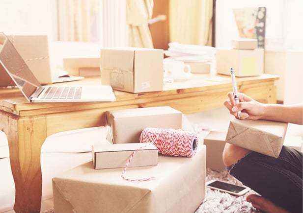 Packed boxes laying around with a laptop on the table and a hand writing with a pen on the box that's saddled on the person's knee.