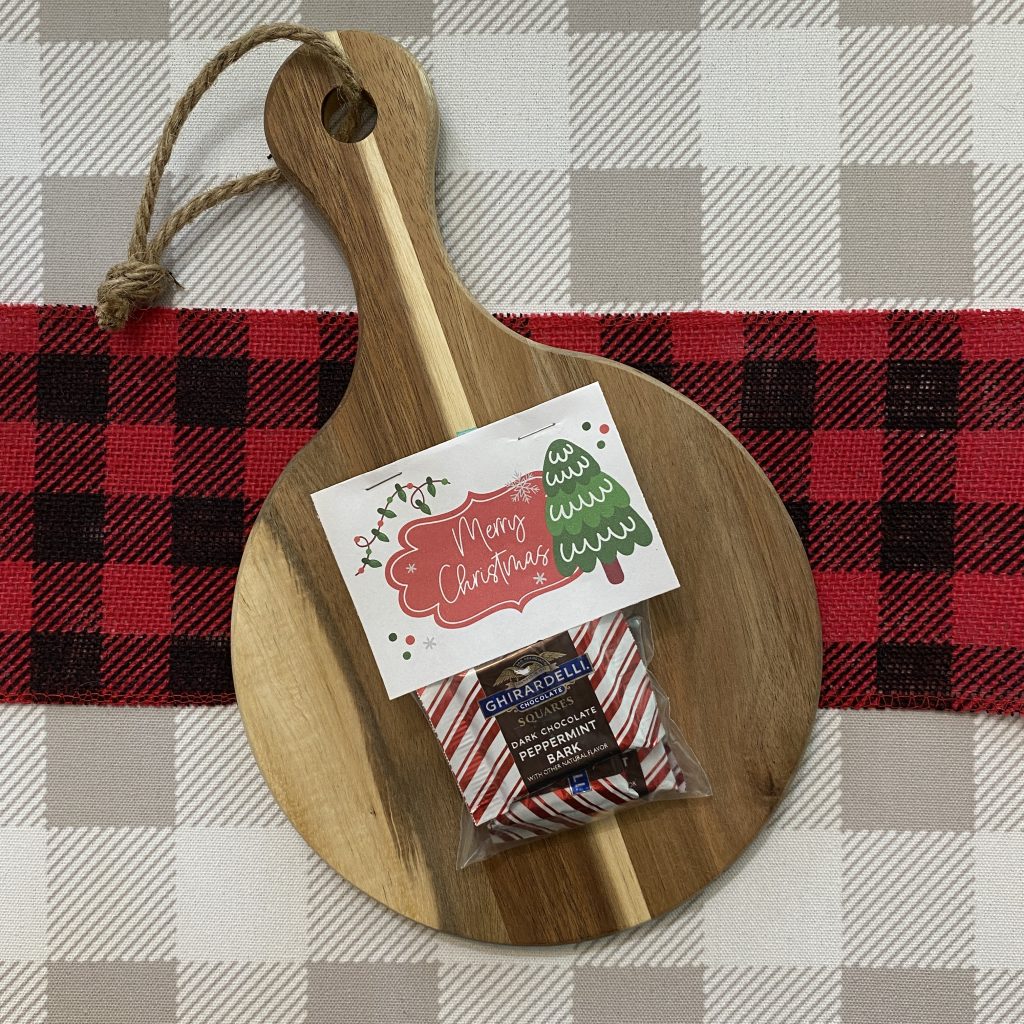 Merry Christmas tag stapled to a treat bag atop a wooden board laid on top of plaid fabric.