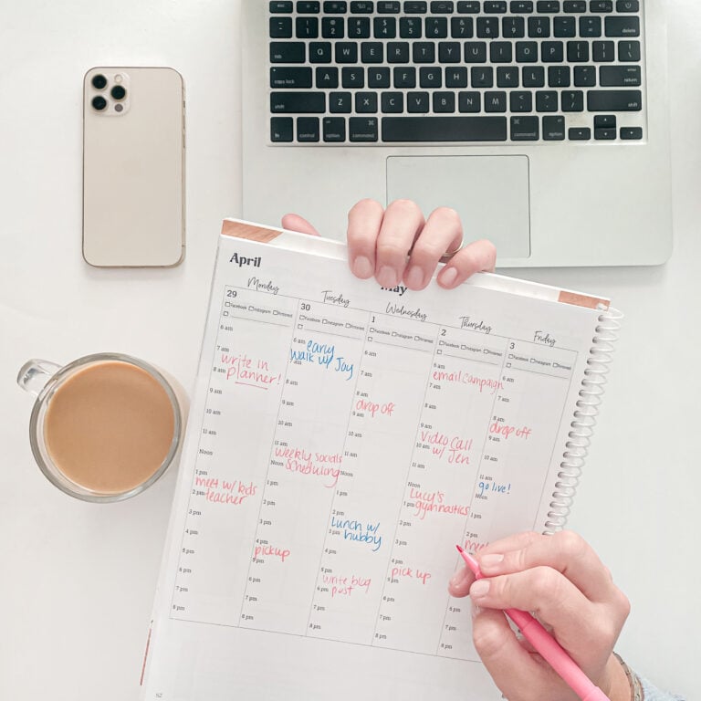 A hand writing on the "Marketing PLanner for Entrepreneurs" planner with a glass of coffee, smartphone, and laptop on the table.