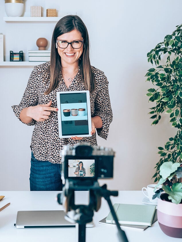 Happy businesswoman having online presentation of modern clay flowerpot and using camera for live broadcast online on the internet.
