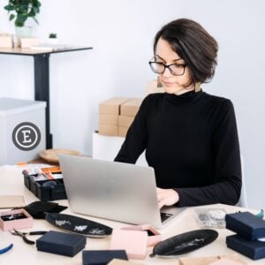 Etsy biz idea with lady at laptop and creative items on her desk.