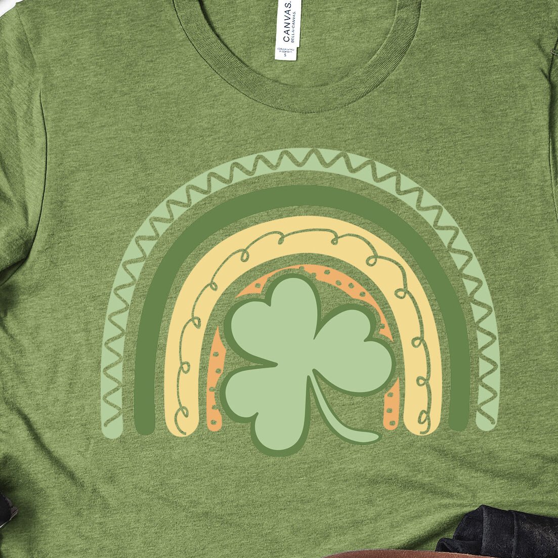 Green T-shirt design featuring a shamrock within a boho rainbow, styled with black shoes and plants. At the bottom right is the We Can Make That logo.